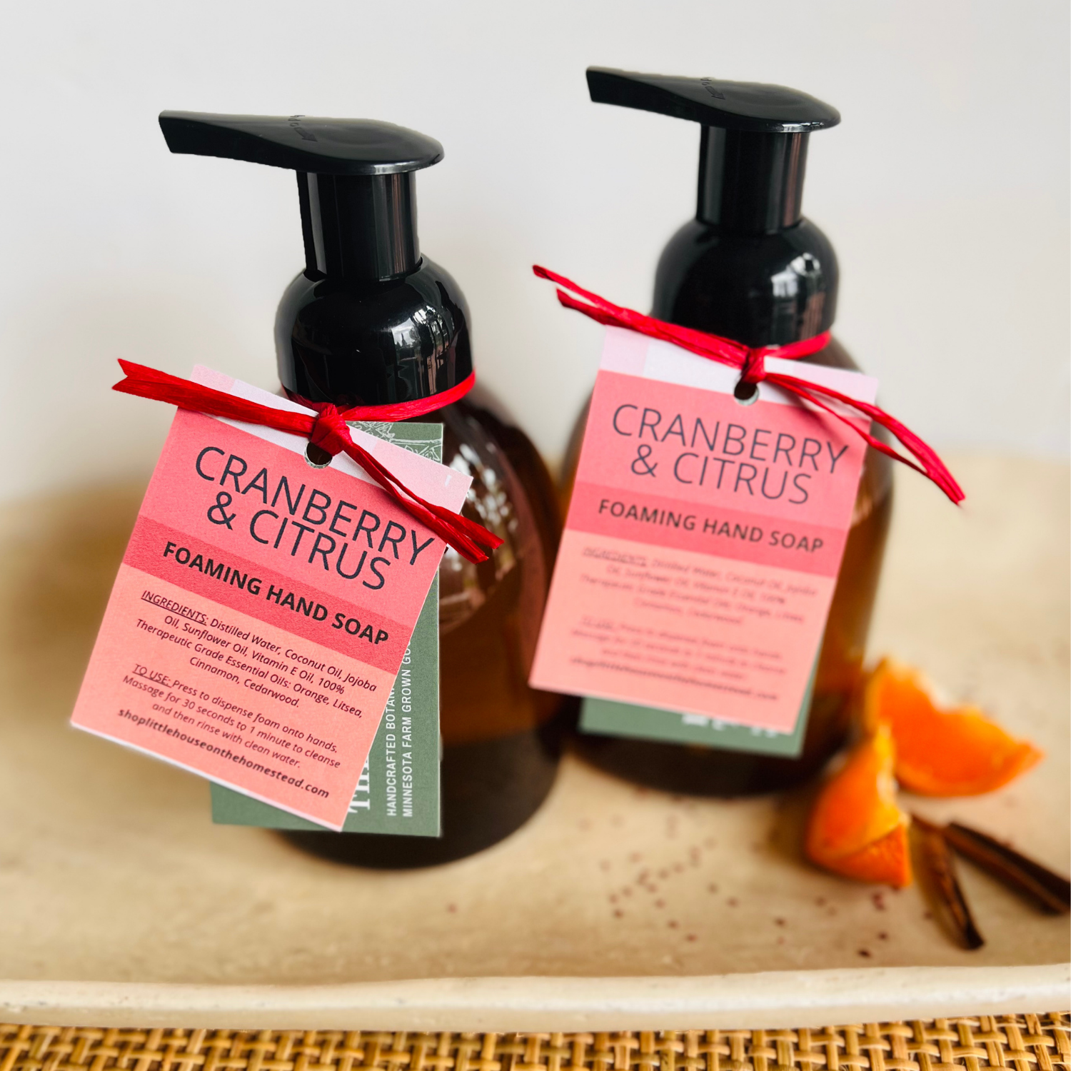 Foaming hand soap bottles styled with orange wedges and cinnamon sticks on a ceramic natural colored tray. Bright red and pink tag with raffia ribbon.